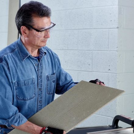 A construction worker wearing a jean jacket holding an angled cut fiber cement board with their right hand and using their left hand to feel the smooth edge of the cut
