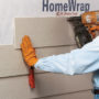 Construction worker holding up a piece of fiber cement Hardie Board with a red Malco FCFG tool while using a nail stapler to staple the board onto the side of a house