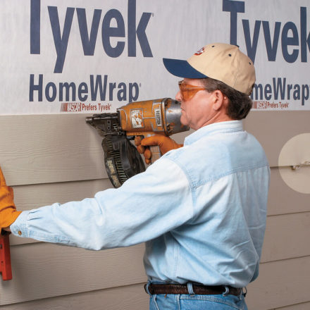 Construction worker using a nail gun to staple a fiber cement siding board on to a house that is being held up with Malco's siding gauge tools