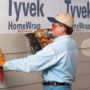 Construction worker using a nail gun to staple a fiber cement siding board on to a house that is being held up with Malco's siding gauge tools