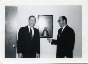 Black and white image of two men standing next to a bell and one of them ringing it
